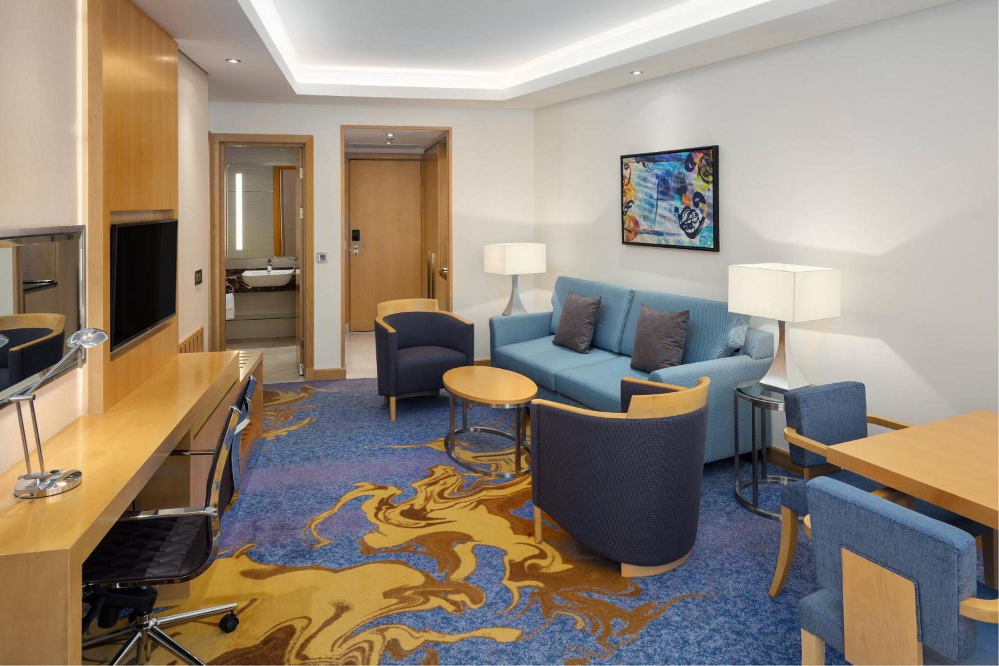 喜来登麦加贾巴尔卡巴酒店 Mecca 外观 照片 The photo displays a modern hotel room or suite. The room features a comfortable seating area with a blue couch and two blue armchairs surrounding a small round table. A wooden desk with a black office chair is positioned against one wall, accompanie