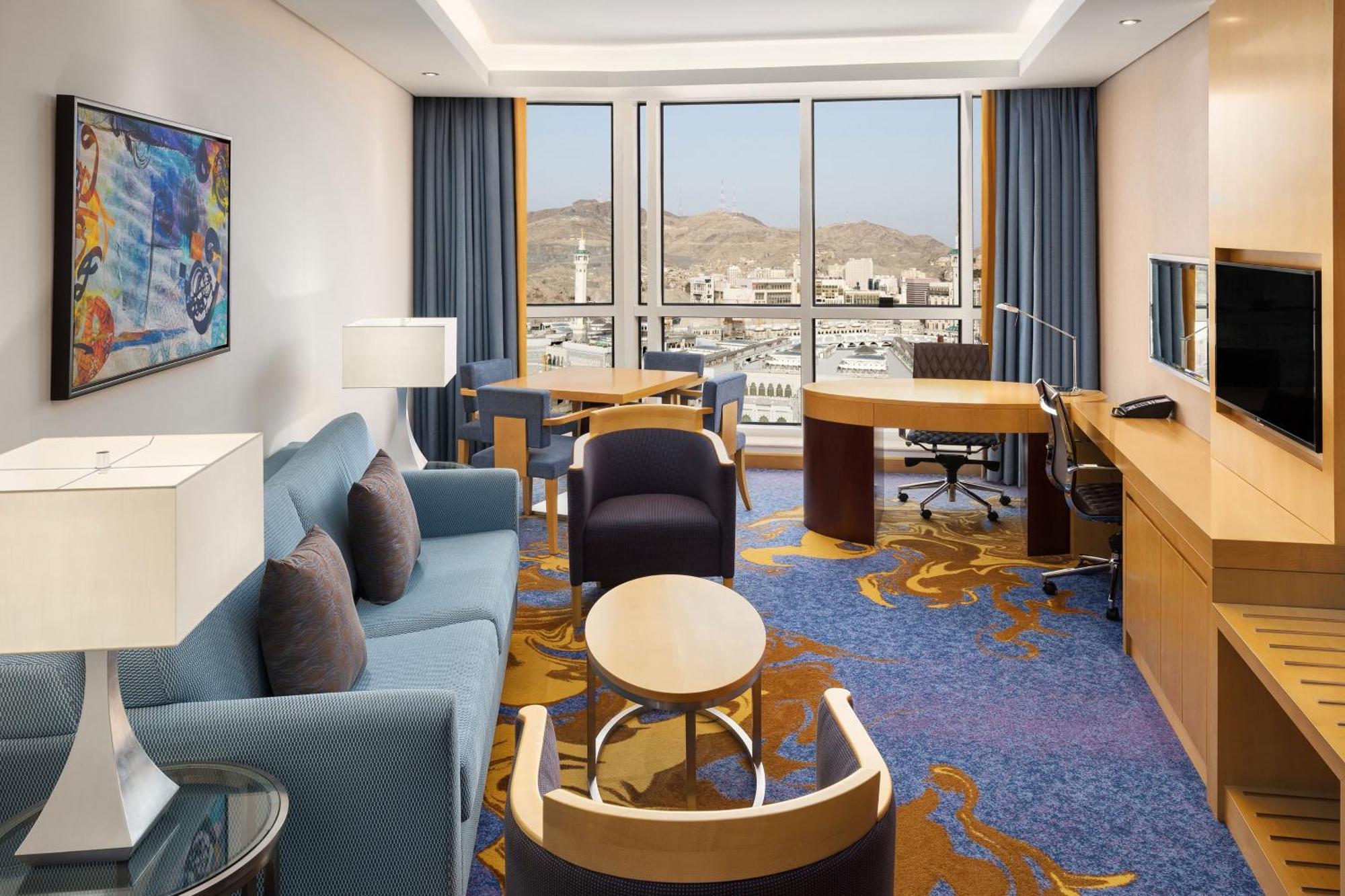 喜来登麦加贾巴尔卡巴酒店 Mecca 外观 照片 The photo shows a well-decorated hotel room featuring a modern and comfortable interior. There is a cozy seating area with a blue sofa and armchairs, complemented by a round coffee table. A desk with a chair is also present, ideal for work. The room 