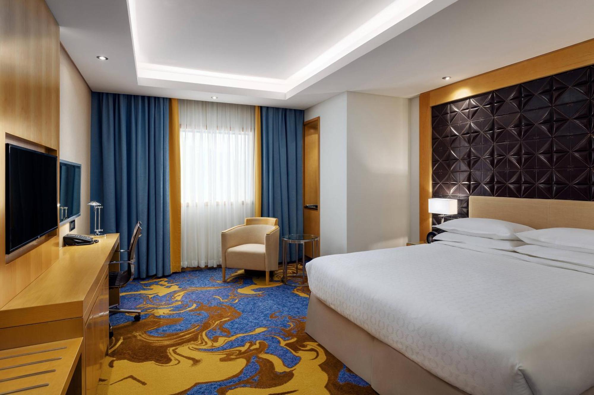 喜来登麦加贾巴尔卡巴酒店 Mecca 外观 照片 The photo shows a modern hotel room featuring a comfortable double bed with a light-colored headboard. There's a large window dressed with blue curtains, allowing natural light to enter. The walls are painted in neutral tones, and one wall has a text